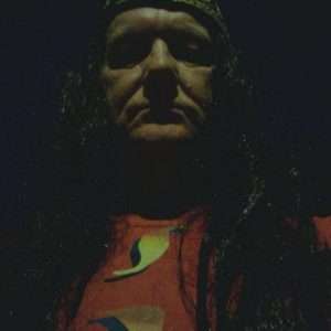 STEViE ZiPP wearing a long dark wig and fezz hat against a dark background